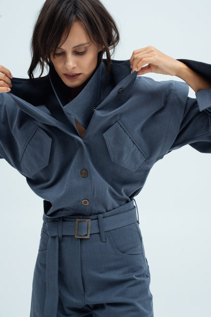 A model wearing a Aisuru jacket in stone blue color  shirt and Rei  trousers, holding the collar open to show the inner collar that can be removable.