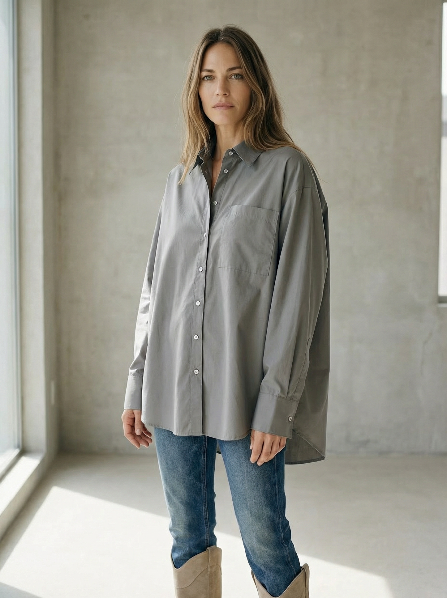 Woman wearing a gray shirt and blue jeans standing in a minimalistic room with concrete walls.