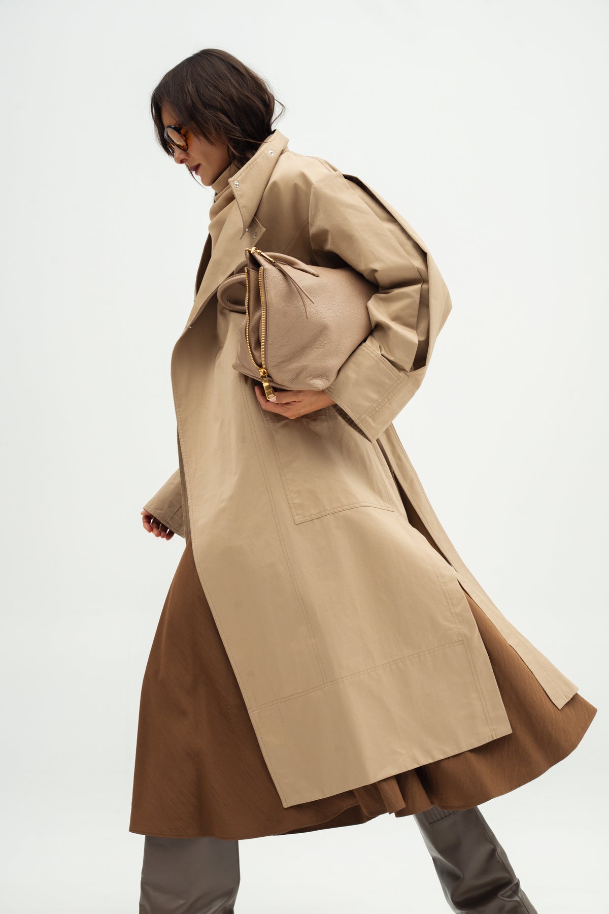A woman wearing Resident stranger  a long Moani trench coat over a Kaya nomad brown skirt, holding a matching beige clutch bag, photographed against a plain light gray studio background.