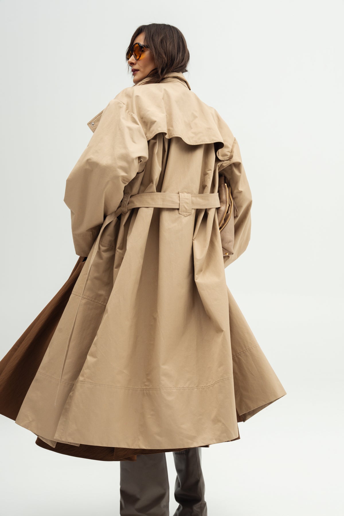 A woman from the back wearing Resident stranger  a long Moani trench coat over a Kaya nomad brown skirt, holding a matching beige clutch bag, photographed against a plain light gray studio background.