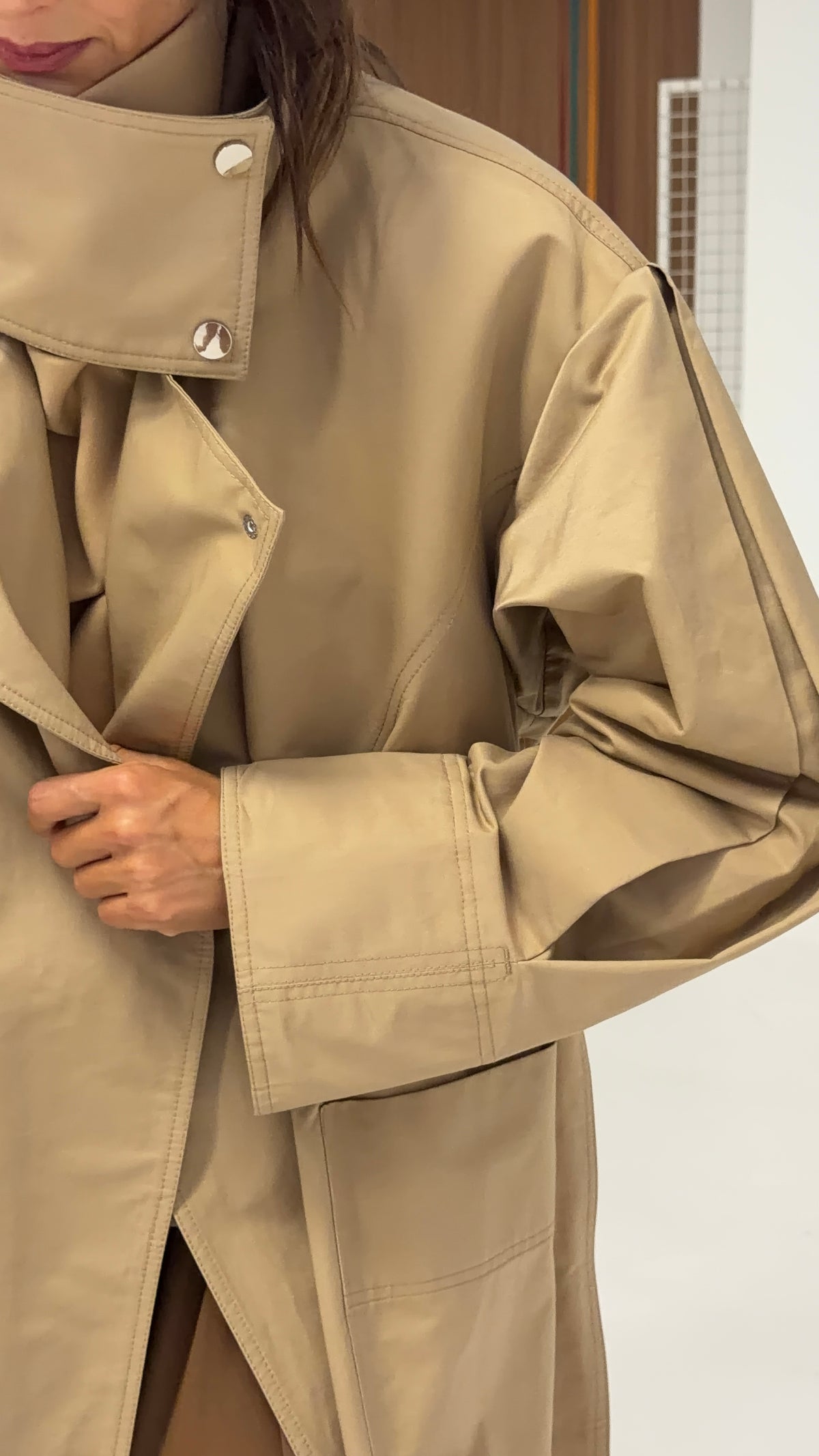 A close-up of a person wearing an oversized Moani trench coat with a high collar and silver-tone snap buttons, shown indoors against a light wall.