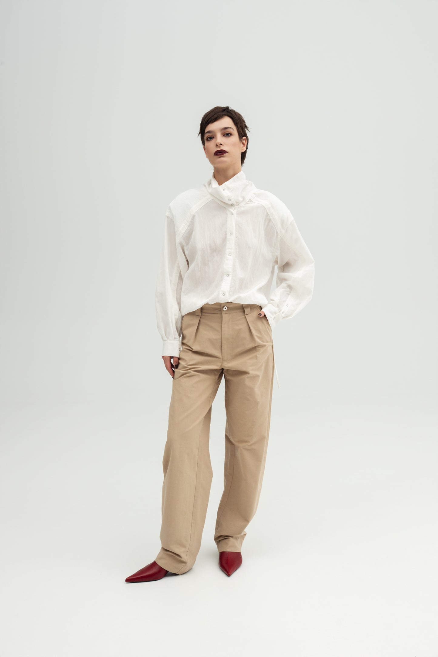 A model wearing a white Nume high-neck sheer long-sleeve blouse Resident Stranger with beige Miso wide-leg trousers Resident Stranger , and dark red pointed shoes.