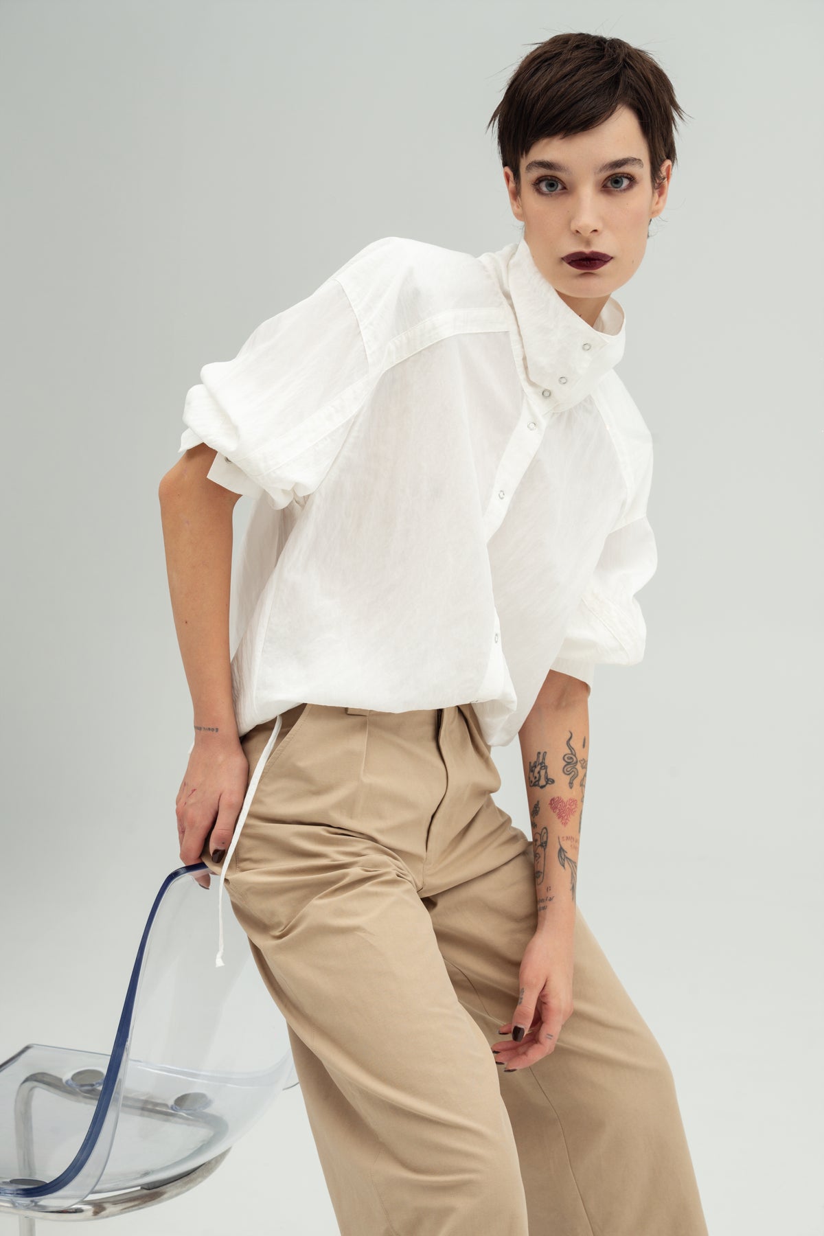 A model wearing a white Nume  button-up shirt Resident Stranger with a high collar,  paired with beige Miso trousers Resident Stranger.