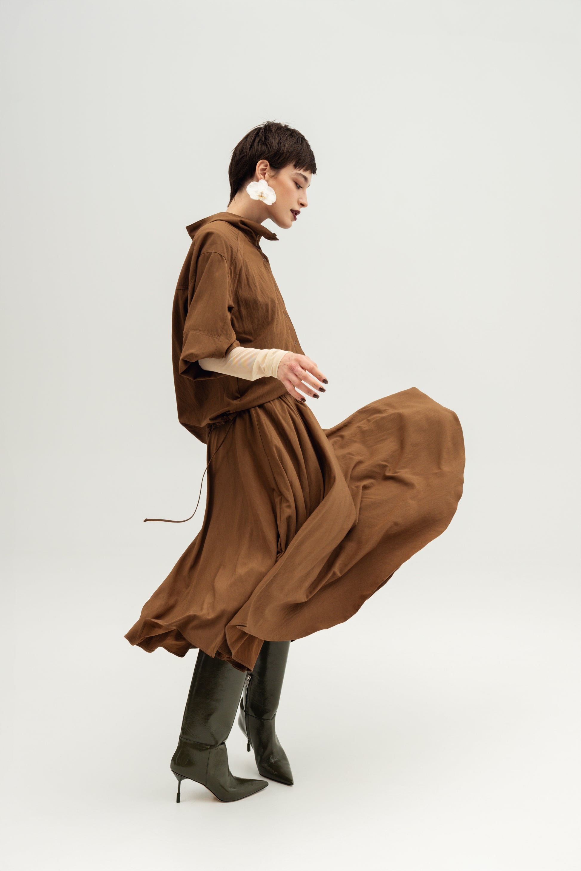 A model wearing a Resident Stranger Nomad brown oversized Nume shirt  with a high collar, round skirt  and dark green pointed-toe boots, captured in motion against a light gray studio background.