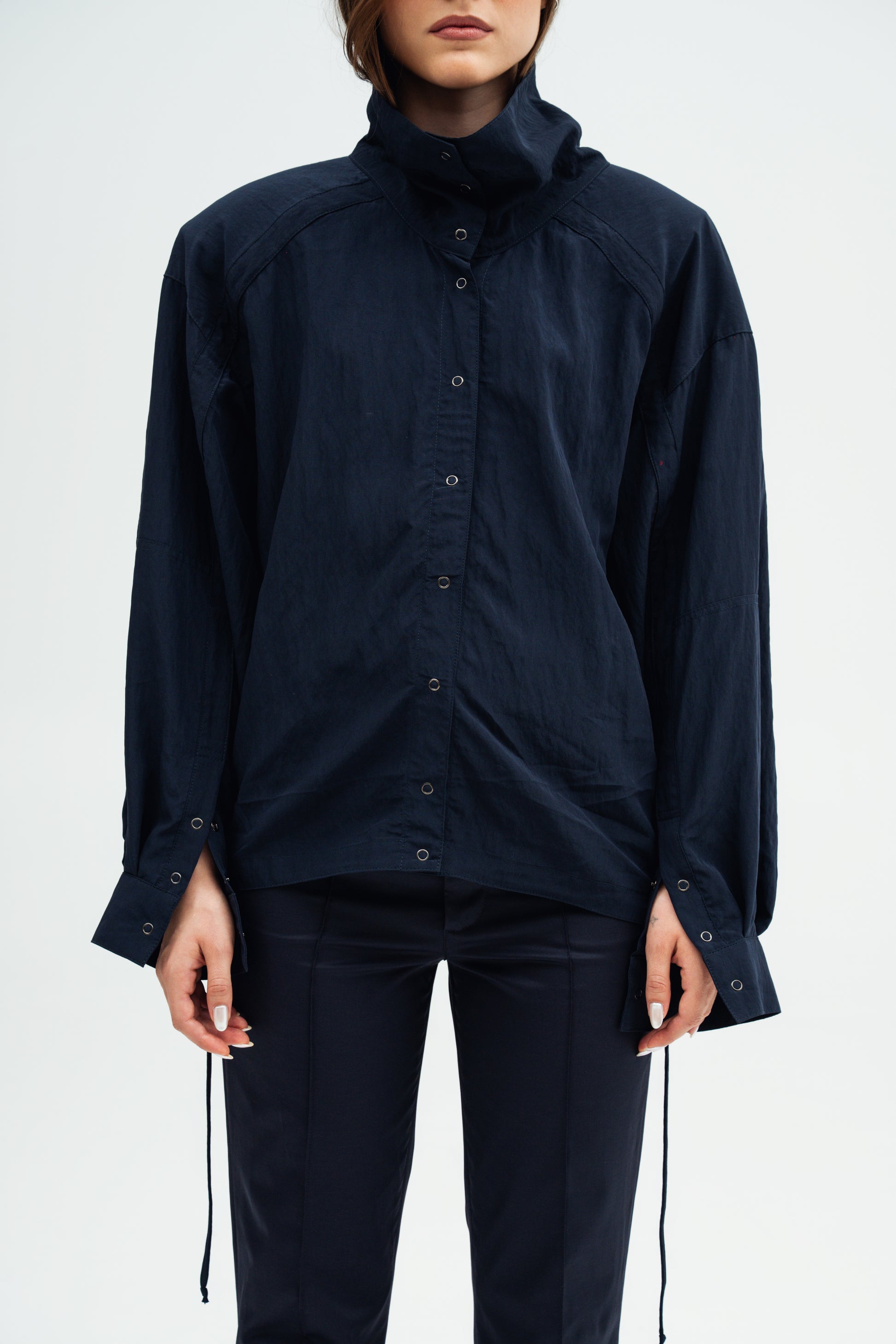 A model wearing a dark navy blue high-neck button-up shirt Nume Resident Stranger  with long sleeves and matching dark pants, shown from the neck down against a plain light background.