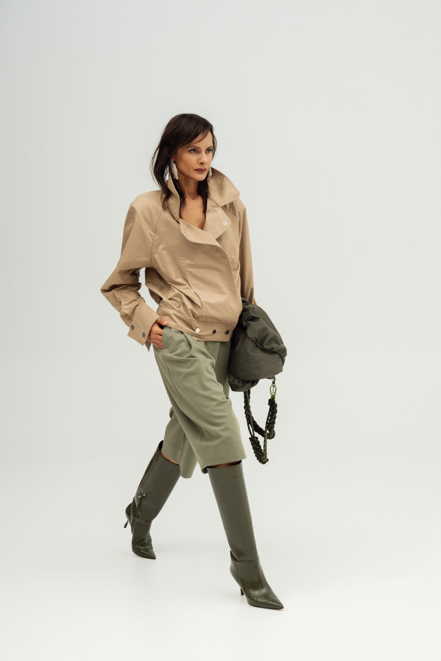 A model is shown from the waist down wearing a Kali oversized jacket, Ukiyoro olivine bermuda , and olive green knee-high boots, while carrying a dark green shoulder bag against a light gray studio background.