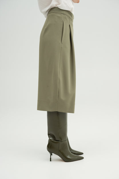 A close-up - side of a model wearing Resident Stranger Ukiyoro bermuda in a olivine color, styled with a white top and dark leather boots against a light gray background.