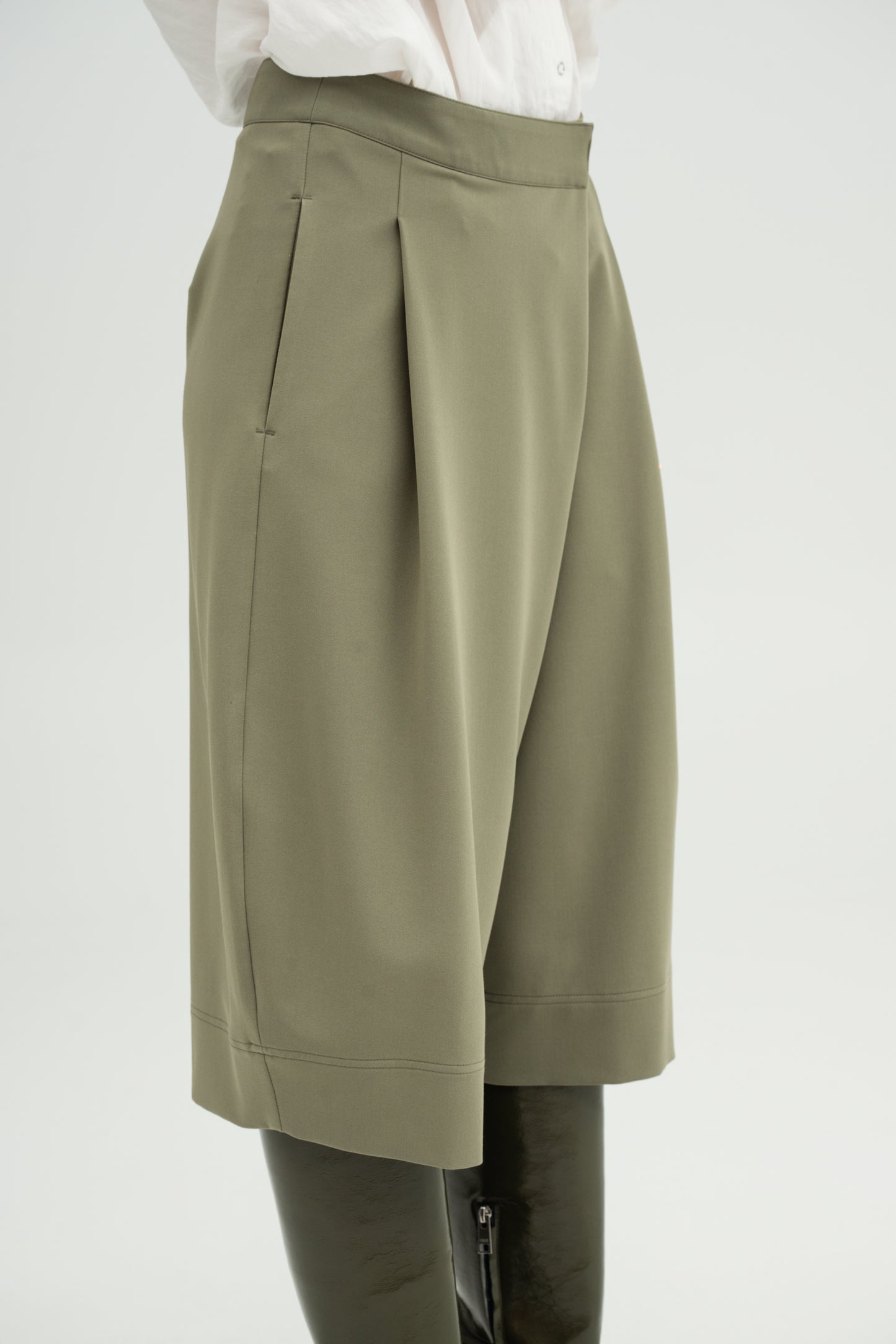 A close-up of a model wearing Resident Stranger Ukiyoro bermuda in a olivine color, styled with a white top and dark leather boots against a light gray background.