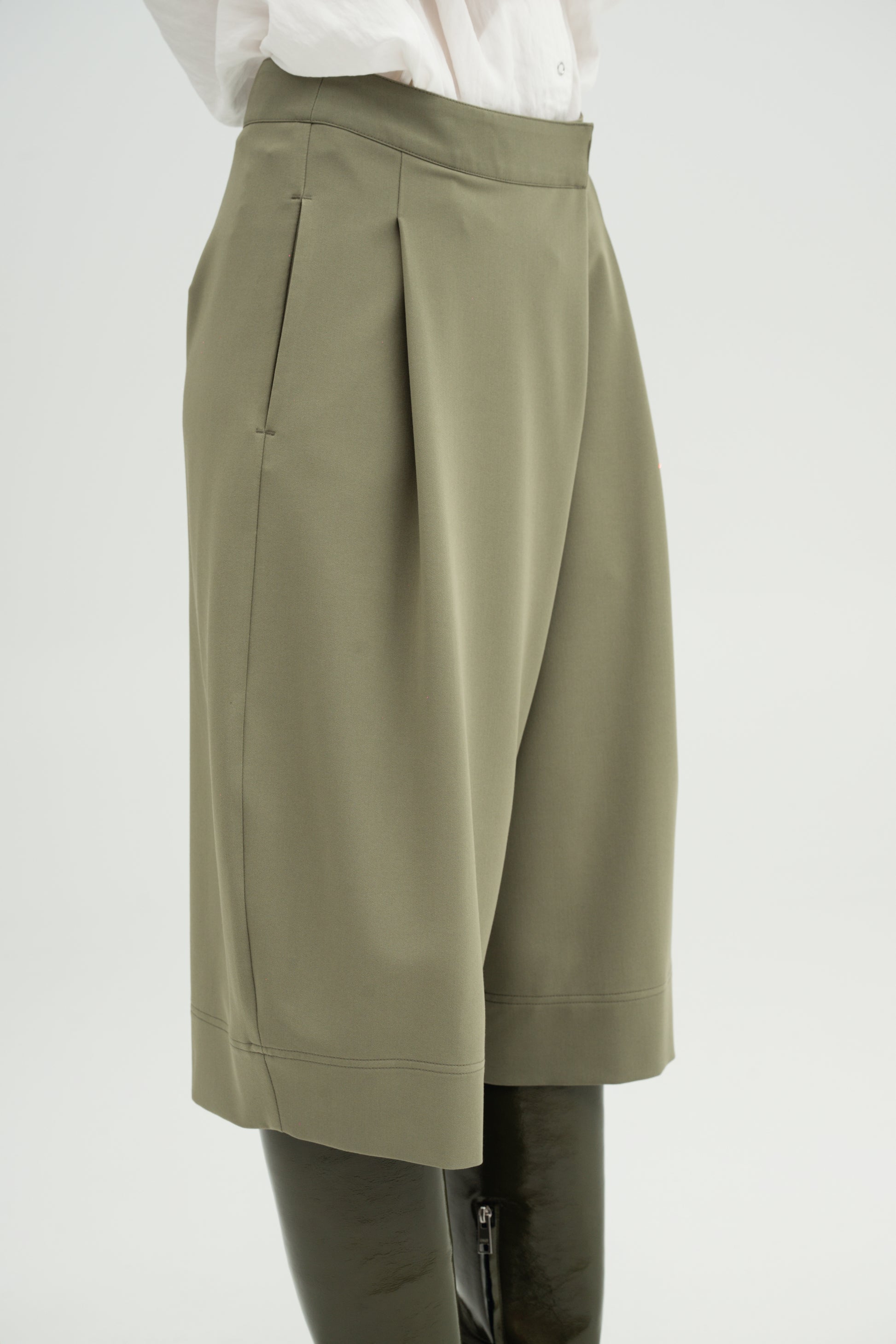 A close-up of a model wearing Resident Stranger Ukiyoro bermuda in a olivine color, styled with a white top and dark leather boots against a light gray background.